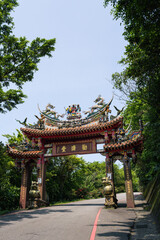 Obraz premium Traditional Chinese archway gate with ornate roof carvings and dragon sculptures on a paved road leading into a green forest park in Taiwan