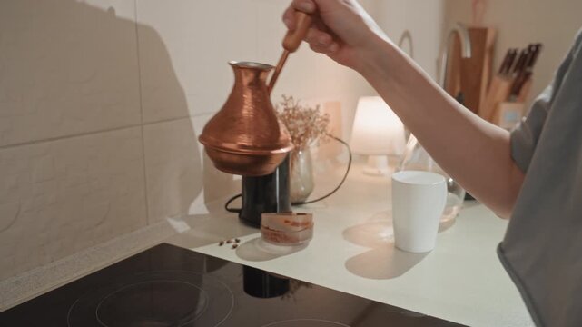 Copper pot used by woman to pour coffee. Ambient setting with warm shadows and textured countertop scene. Home barista woman gently pouring hot coffee from copper vessel into mug with care
