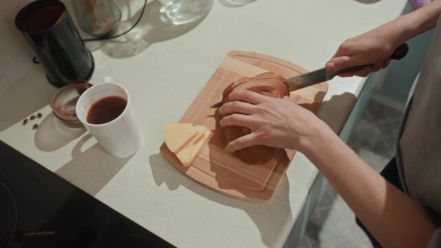 Kitchen counter hands slicing bread with coffee mug and cheese, home cook gently cuts rustic loaf in warm morning light, intimate cozy atmosphere suggests music lover taking slow breakfast moment,