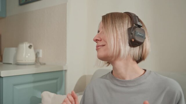 Woman headphones dancing on couch eyes closed, rhythmic hand gestures, smiling expression, kitchen backdrop, mug on side table, immersive listening, upbeat tempo, private concert vibe, energetic