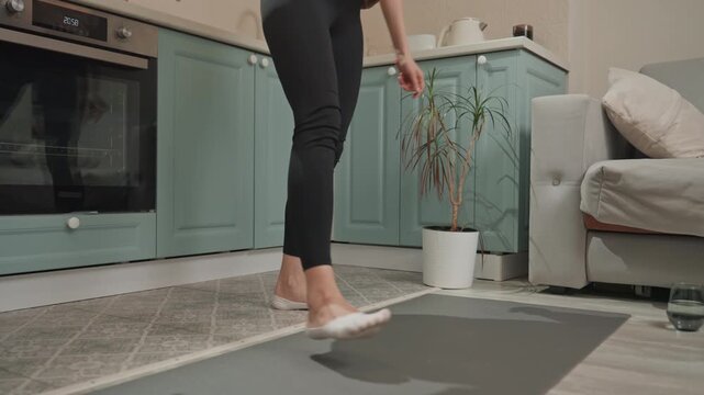 White woman in kitchen unrolling mat before practice, headphones around neck, casual hoodie, barefoot on rug, preparing for home movement session with upbeat music and focused balance.