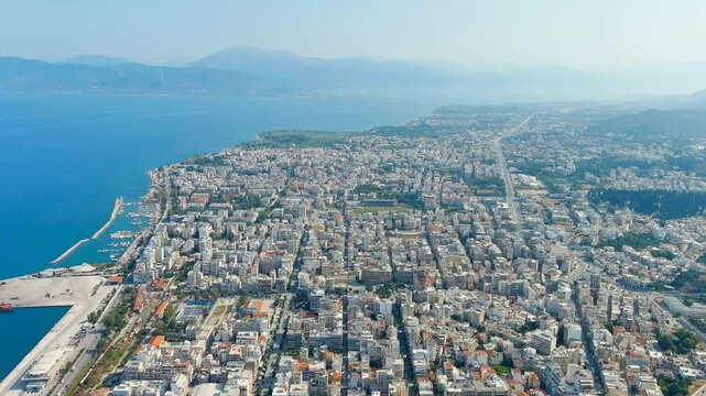 Patras, Greece. the central city and working port along the Gulf of Patras, white buildings and terracotta roofs under bright summer sun, with the Rio-Antirrio Bridge visible on the distant horizon. 
