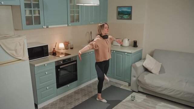 Caucasian woman dancing in kitchen to music with headphones, spinning and stepping across rug, teal cabinets and sofa backdrop, lively expression and upbeat tempo street dancer energy meets home