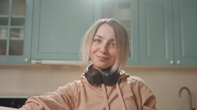 White woman raising arms with headphones kitchen, ecstatic joyful release with flowing gestures and radiant smile, casual hoodie, warm backlighting, spontaneous home performance full of bliss