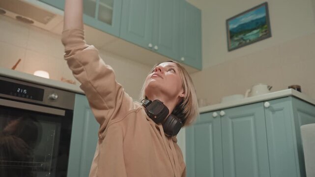 White woman reaching toward upper cabinet, headphones resting on neck, expressive upward stretch and subtle dance moves, playful mood, teal kitchen, painting on wall, dynamic extension for energy