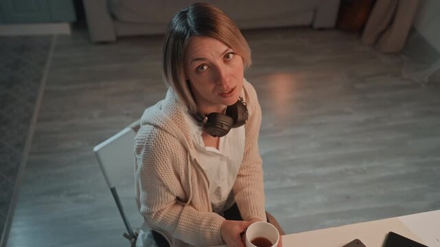White woman with headphones gesturing, desk scene with coffee cup, expressive hand movements, vlogger or podcaster rehearsing lines, playful banter and occasional sip, candid smile, evening glow,