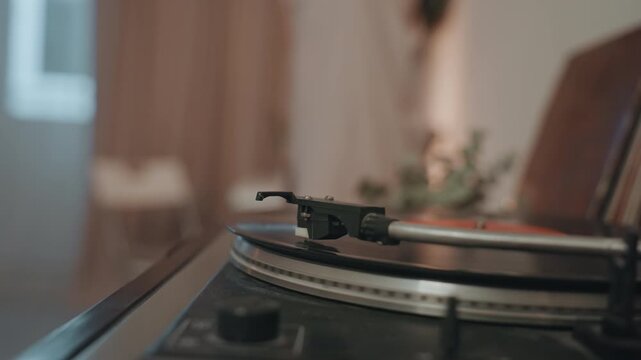 Turntable stylus poised over vinyl, blurred figure moving in background, soft lamp glow, close focus on needle tip, quiet ritual of music lover preparing playback, vintage tones, shallow depth,
