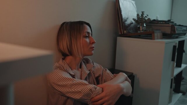 White woman leaning against wall near cabinet with turntable striped shirt soft orange and teal lighting reflective gaze record listener reminiscing intimate home scene melancholic calm