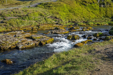 A flowing stream cascades over moss-covered rocks in a lush green valley.,ICELAND © SHUNJI ZHOU