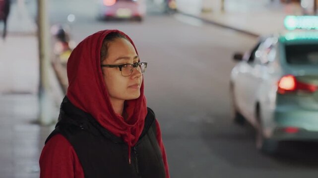 Nighttime street pedestrian. Person in red pauses watching traffic. Hooded individual observes passing vehicles at night. Redclad pedestrian glances attentively at moving cars during evening