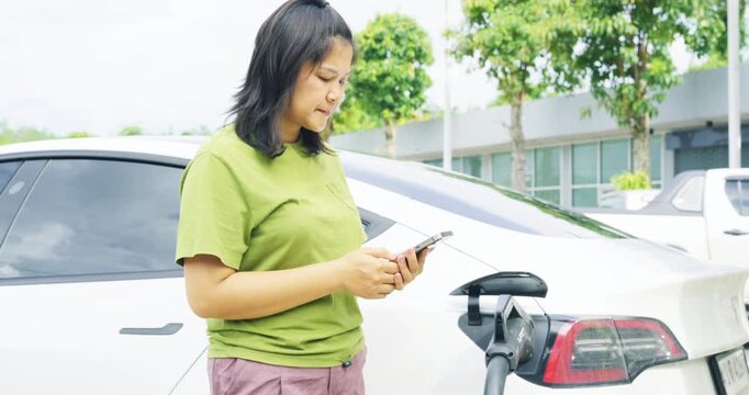 Casual woman use outdoor station EV car charge with mobile phone transport technology