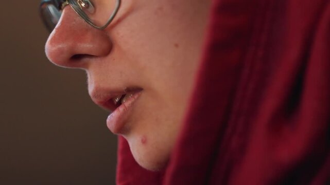 Night closeup of intimate speaker in red hoodie and glasses, lowlight mood with warm tones and subtle shadow. Quiet confession or storytelling moment, soft textures on fabric, focused lips and breath,