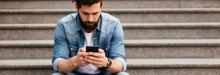 Obraz premium Bearded man in denim shirt sits on concrete stpic outdoors. He holds a smartphone and looks at the screen, typing or browsing. He seems focused on his mobile device.
