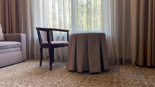 Interior of an elegant hotel room with a wooden chair and a round table by the window with curtains