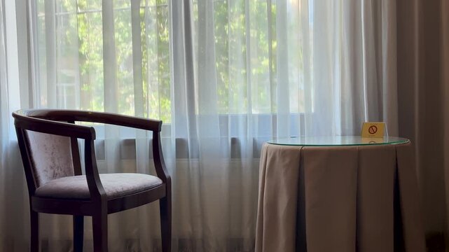 Interior of a cozy hotel room with a wooden chair and a small round table with a no smoking sign by the window with white curtains