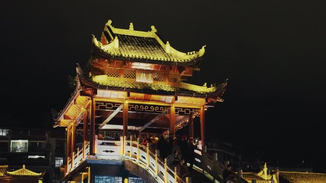 Lit pagoda bridge night walking atmosphere, warm golden lanterns outlining ornate rooflines and curved staircase, empty promenade and quiet urban skyline, moody dark sky framing traditional