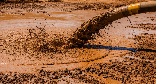 Industrial sludge pumping from a pipe onto muddy ground. Brown liquid waste splashing during a mining or construction project. Waste management concept for industrial marketing.