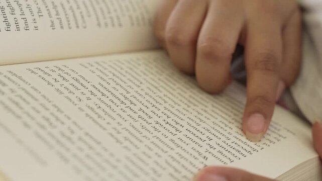 Close-up of a woman's hand gently pressing and smoothing an open book page while reading, with clearly visible printed text and soft natural window light illuminating.