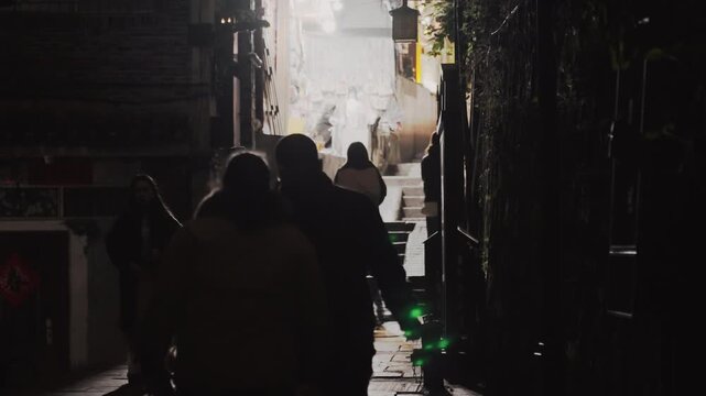 Night walking through narrow wet alley with silhouetted group moving toward bright exit, backlit figures and lamplight casting reflections on puddled pavement, commuters and shoppers in coats