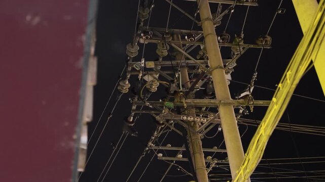Night utility pole with tangled cables, lamp glow, transformer hardware, intersecting wires, urban skyline edge, quiet moody atmosphere, industrial texture, maintenance perspective, cinematic