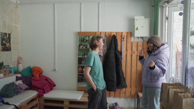 Pottery workshop interior seller helping customer. Cozy studio with wooden pegboard of coats, bench seating, stacked parcels and window light. Person in green shirt guides visitor in purple coat