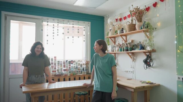 Two women in pottery workshop arranging shelves and chatting, sunlit studio filled with ceramic jars, potted plants, wooden table, tools and aprons one inspects glaze bottles while other laughs, cozy