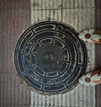 Gata de Gorgos, SPAIN - May 20, 2026.Vintage cast iron manhole cover on red and white striped sidewalk with sandals during summer day