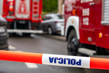 A red and white police warning tape with the Polish word POLICJA in the foreground, blocking a street at an accident scene with blurred red fire trucks and a damaged silver car in the background. © Adam