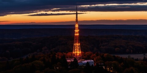 Obraz premium Glowing comms tower lights up forest at dusk. Aerial view shows rural area with trees and hills. Evening light creates warm glow on structure and landscape. Wireless signal reaches remote locations.