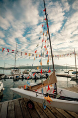 Obraz premium Little sailboat docked at a wooden jetty with vibrant pennant flags, children sailing camp adventure concept.