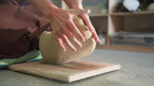 Caucasian apprentice centering clay in workshop, hands pressing sides and top to form symmetrical ball, gritty studio floor, nearby shelves with glazes and tools, focused training moment showing
