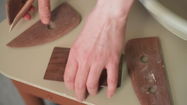 Closeup pottery workshop tool selection and arranging on wheel shelf hands pick ribs sponge and template, checking fit and balance intimate studio focus with textured wood and clay dust