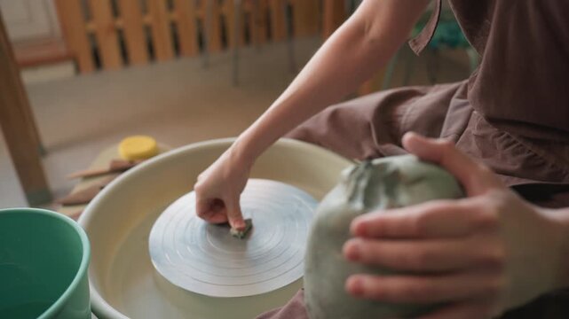 Closeup hands shaping clay on wheel, white apprentice centers lump with gentle pressure, sponge and bucket at side, soft natural light, tactile texture and focused rhythm in small studio