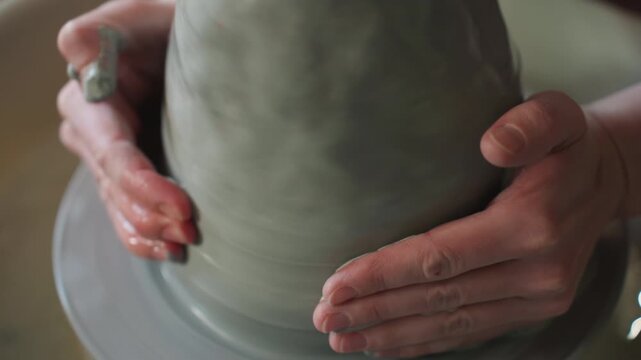 Craftsman shapes wet clay on spinning wheel. Closeup of hands forming vessel from soft gray material. Meditative pottery session where palms gently shape wet clay spinning on wheel