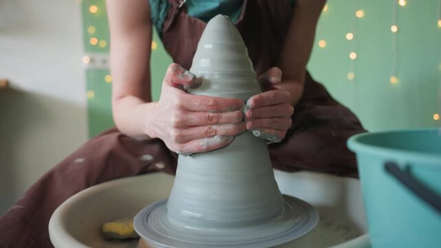 Potter crafting vase. Woman shapes round ceramic vessel. Artist molds coneshaped pottery on spinning wheel. Female artist carefully forms conical clay vessel with bright background lights