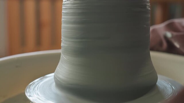 Gentle movements create smooth ceramic surfaces. Craftsman expertly forms slender walls on spinning pottery wheel. Soft studio lighting highlights rhythmic motion of shaping fine ceramic vessels