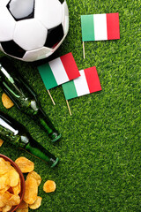 Naklejka premium A soccer ball, green beer bottles, potato chips and Italy flags on green grass create the perfect game viewing atmosphere for Italian fans. Top view with copy space.