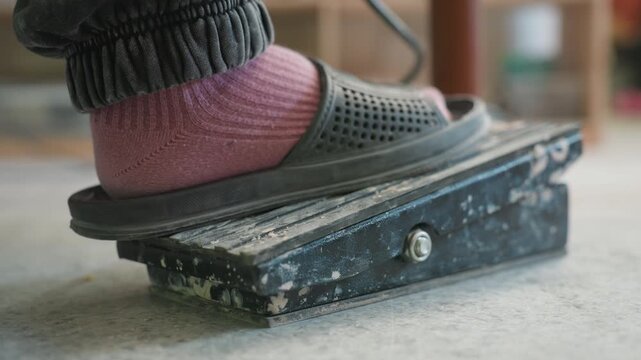 Pottery pedal foot closeup in workshop. Ceramicist presses aged metal pedal to control wheel speed, pink sock visible under black sandal, paintsplattered surface and concrete floor, intimate studio