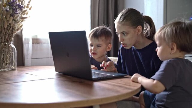 Adult monitors kids studying independently. Caretaker oversees two children engaged in learning. Caregiver attentively observes two children as they focus on their online lessons at kitchen table