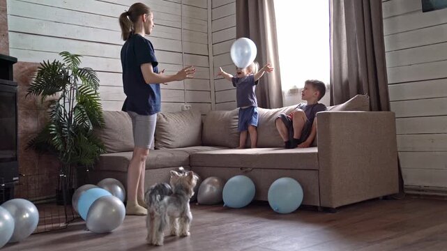 Children playing reflex game with balloons, older sibling guides little ones to catch floating silver balloon while terrier watches, scattered blue balloons on hardwood floor and bright window light