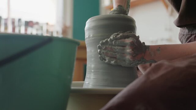 Clay shaping demonstration in welllit studio. Instructor exhibits pottery technique using tall jar near window. Glass shelf illuminated by sunlight in studio where artist molds clay on wheel