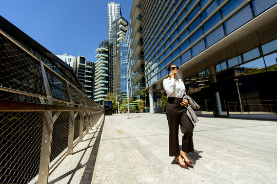 Woman walking in city wearing sunglasses while talking on phone near modern buildings
