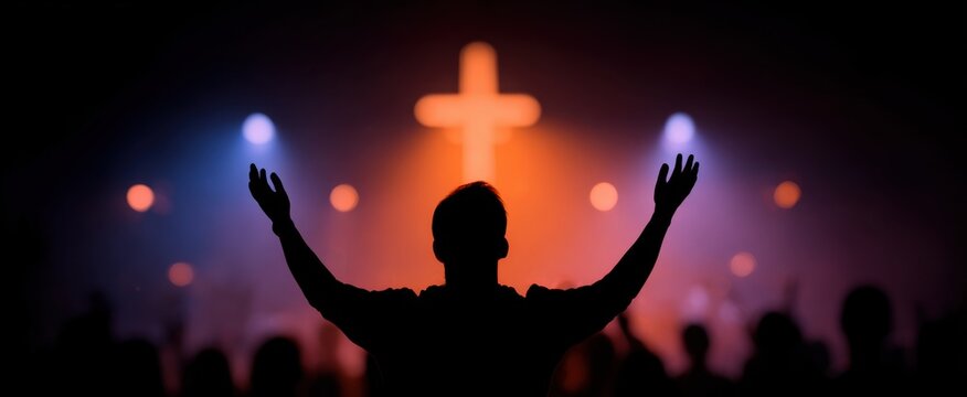Man raising hands in praise at gospel concert with crowd worshiping under bright cross.
