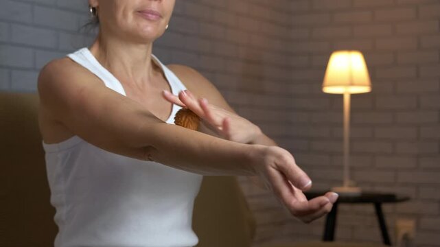 Woman performing self-massage on arm with ball. Mature woman sitting at home in the evening using a small spiky ball to perform a therapeutic self-massage on her arm, relieving muscle tension