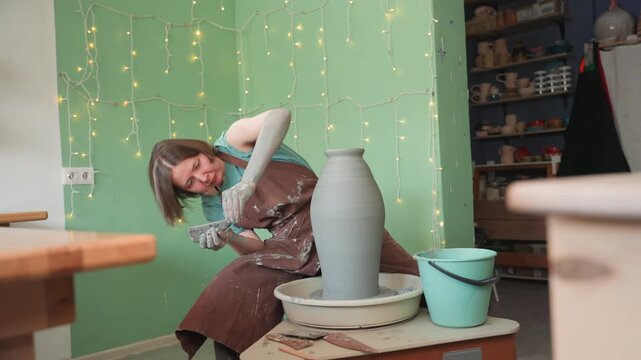 White female potter shaping tall vase on spinning wheel, cozy workshop with string lights and turquoise bucket, brown apron and claysplattered hands smoothing wet surface, slow handmade process