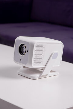 Modern White Portable Home Projector on Stand with Lens Close-Up in Minimalist Interior Setting. 4K Wifi Bluetooth Smart Projector with an adjustable stand for home cinema, presentation or job meeting