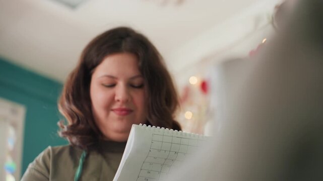 White woman shaping clay on wheel, pottery instructor reading notes while guiding student, hands covered in wet clay, cozy workshop studio with teal wall and soft natural light, notebook checklist