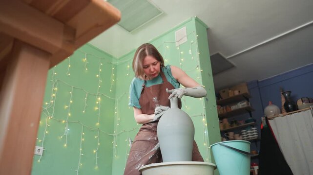 White woman refining vase rim on wheel in tidy pottery workshop, closeup hands trimming clay edge with steady focus, green backdrop, bucket and tools on bench, soft studio light highlighting wet