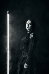 Obraz premium Gothic young woman with long dark hair and black makeup posing by vertical light tube in smoke