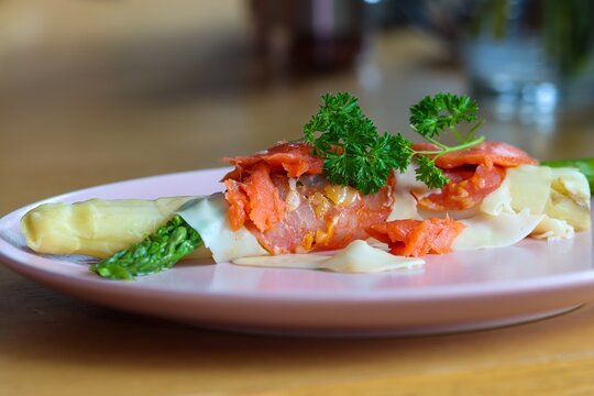 Green and white cooked asparagus wrapped in a piece of ham with cheese, a piece of salmon and parsley on a plate.